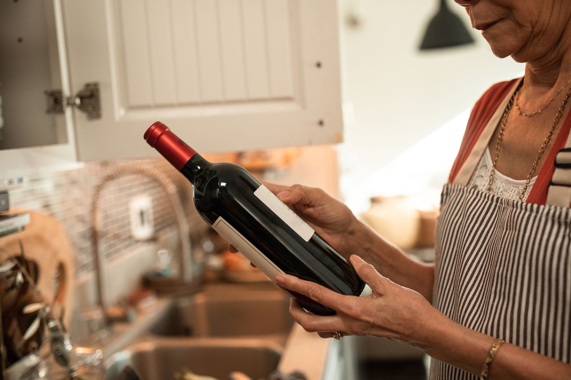 Jak używać resztek wina z Bordeaux w kuchni francuskiej na co dzień Starsza kobieta w fartuchu ogląda butelkę wina w przytulnej kuchni